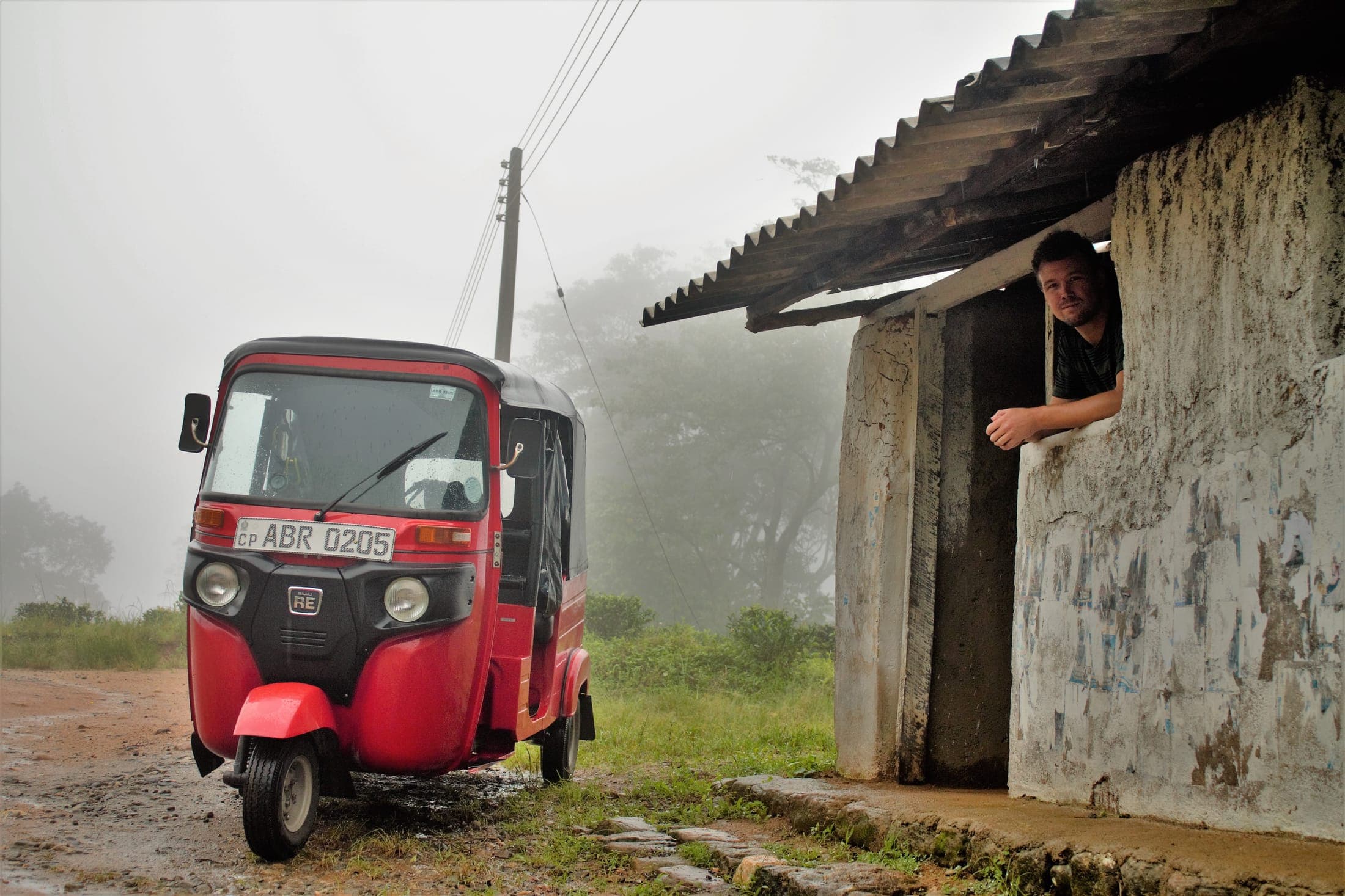 A red tuk-tuk outside a stone hut on a misty mountain road